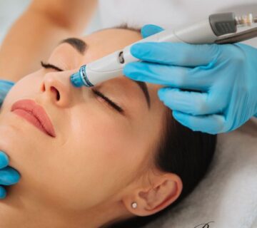 A woman receives a facial treatment with a handheld device while lying down, performed by a professional wearing blue gloves.